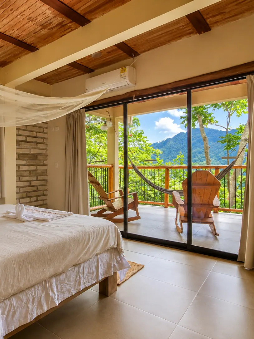 Bedroom with a large white bed and glass doors open to a terrace with a hammock, wooden chairs and green mountains